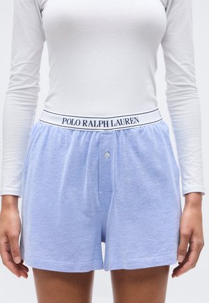 Femme portant un short bleu clair texturé avec une ceinture élastique blanche marquée "Polo Ralph Lauren" et un haut blanc ajusté à manches longues.