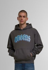 Grå hettegenser med en blå "QUEENS" grafikk. Har en kengurulomme og snor i hetta. Stoffet synes mykt og teksturert.