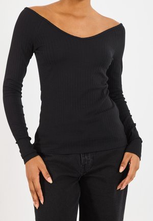 Vrouw draagt een aansluitende zwarte geribde top met lange mouwen en een off-shoulder halslijn en zwarte broek tegen een witte achtergrond.