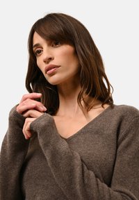 Brauner, weicher Strickpullover mit V-Ausschnitt und langen Ärmeln, der eine strukturierte Oberfläche und gerippte Bündchen aufweist. Das Modell posiert mit gefalteten Händen.