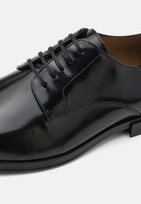 Chaussure habillée en cuir noir avec une finition brillante, comportant cinq œillets et de fines lacets noirs. Coutures accentuées notables le long des côtés.