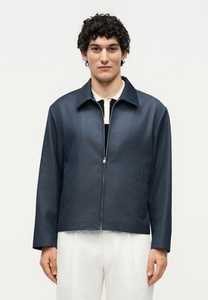 Άνδρας με σγουρά μαύρα μαλλιά φοράει σκούρο μπλε μπουφάν με φερμουάρ πάνω από λευκό πουκάμισο και λευκό παντελόνι, στέκεται σε απλό φωτεινό φόντο.