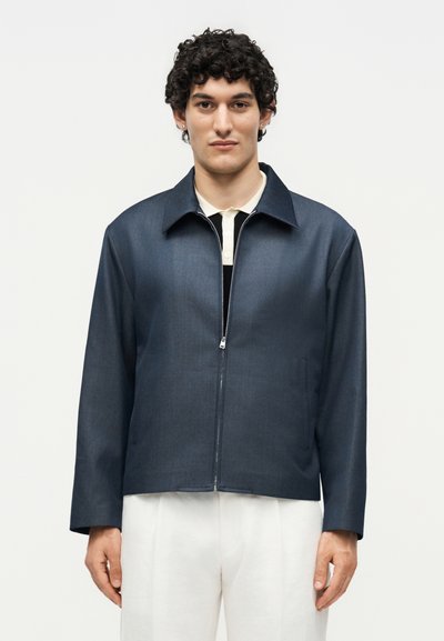 Homme aux cheveux noirs bouclés portant une veste bleu foncé zippée par-dessus une chemise blanche et un pantalon blanc, debout devant un fond clair uni.