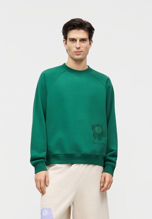 Mies, joka on pukeutunut vihreään pyöreäkauluksiseen collegepaitaan, jossa on hienovarainen kukkalogo helman lähellä, ja vaaleanbeigeihin housuihin, joissa on purppuranvärinen kukkakuosi.