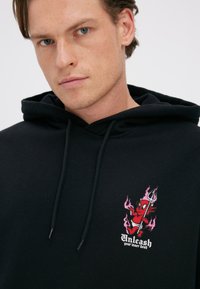 Ung mand iført sort hættetrøje med lille rød djævel-tegneserie, der holder en trefork og flammer, tekst "Slip din indre djævel løs" på brystet.