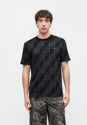 Mladý muž stojící, oblečený v černém tričku Adidas s diagonálním vzorem loga a tmavých maskáčových cargo kalhotách na jednoduchém pozadí.