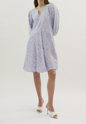 Kobieta w jasnoniebieskiej sukience do kolan z abstrakcyjnym złotym wzorem i bufkami na rękawach, w połączeniu z białymi szpilkami wykończonymi futerkiem.