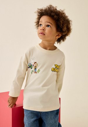 Lille barn med krøllet hår iført en beige langærmet Toy Story-trøje og blå jeans, kigger opad og står ved siden af en rød blok.