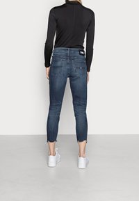 Jeans in denim scuro con un fit accorciato e affusolato, dotati di un orlo con bordi grezzi. Le tasche posteriori e l'etichetta del logo sulla vita sono visibili.