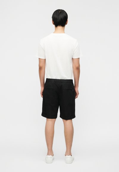 Μαύρο σορτς με διπλωμένο τελείωμα, συνδυασμένο με απλό λευκό μπλουζάκι. Η εμφάνιση ολοκληρώνεται με λευκά αθλητικά παπούτσια. Εμφανίζεται η πίσω όψη.