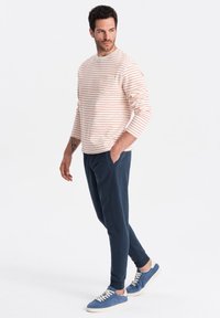 Gestreepte lange mouwen shirt in lichtcrème met rode strepen, gecombineerd met marineblauwe joggingbroeken en blauwe suède sneakers, die een casual sportieve look laat zien.