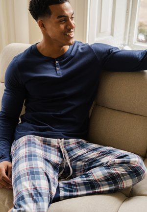 Chemise henley à manches longues bleu marine avec trois boutons, associée à un pantalon de pyjama à carreaux à cordon de serrage dans des tons de bleu, blanc et rouge, assise sur un canapé beige.
