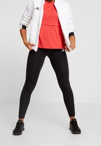 Bílá zipová bunda, červený sportovní top a černé legíny. Obsahuje loga Adidas, přiléhavý střih a hladkou texturu po celé ploše.