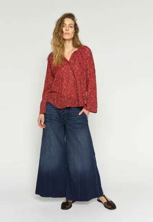 Femeie cu păr lung, ondulat, purtând o bluză roșie cu imprimeu floral, blugi largi de culoare albastru închis și pantofi negri țesături, stând în picioare pe un fundal alb simplu.