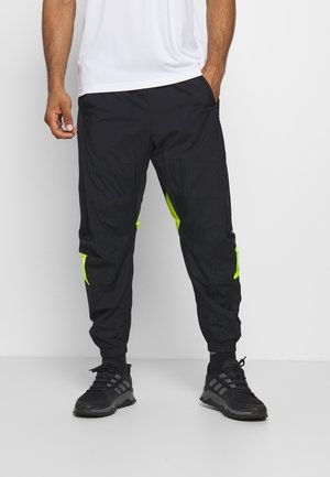 Homme portant un pantalon de jogging noir avec des accents jaune fluo, des chaussures de sport noires et une chemise blanche, debout les mains dans les poches.
