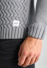 Szary dziany sweter z fakturowanym wzorem w rybią ość, z prążkowanymi mankietami oraz kwadratową metką w dolnym lewym rogu z napisem "ONLY & SONS".