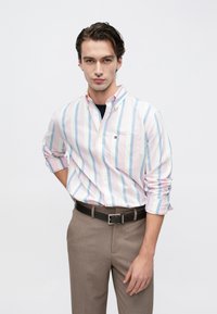 Πουκάμισο με κουμπιά σε παστέλ ροζ και μπλε ρίγες, μακριά μανίκια γυρισμένα. Σκούρα ζώνη, ανοιχτό καφέ παντελόνι, με διακριτικό λογότυπο μάρκας στην τσέπη.