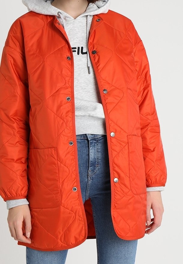 Kurtka pikowana w kolorze pomarańczowym z zapięciem na napy, dwoma przednimi kieszeniami i luźnym krojem. Nosi ją w zestawieniu z szarym kapturem i niebieskimi dżinsami.