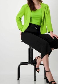 Blazer texturizado verde brillante sobre una blusa a juego; pantalones negros cortos con cinturón y sandalias de tacón negro brillantes.
