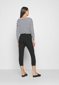 Femme avec de longs cheveux bruns portant un pantalon noir court, une chemise à manches longues rayée noir et blanc, des boucles d'oreilles créoles et des chaussures noires à enfiler.