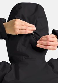 Veste noire imperméable avec une capuche ajustée. Des mains ajustant un cordon, mettant en valeur une texture lisse et mate ainsi que des détails de conception fonctionnels.