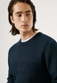 Jūrinis megztinis su tekstūruota paviršiumi ir subtiliu logotipo akcentu priekyje. Rūbų apykaklė ir rankogaliai su briaunomis; vidutinio storio.