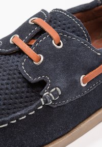 Mörkblå mockaskor med perforerat mönster, bruna lädersnören, silverögon och kontrasterande sömmar på en gummisula.