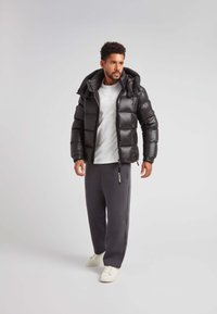 Giacca a vento nera con finitura lucida, chiusura con zipper e cappuccio. Abbinata a pantaloni della tuta grigi e sneakers bianche. Design minimalista.