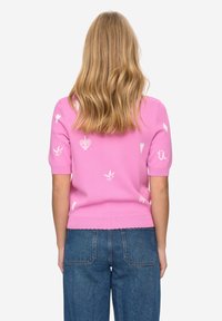 Krátký růžový pletený svetr s krátkými rukávy, zdobený bílými vzory srdcí a květin, s vroubkovaným lemem. Nošený s modrými džínami.