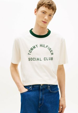 Junger Mann trägt ein weißes T-Shirt mit grünem Kragen und dem Schriftzug "Tommy Hilfiger Social Club", kombiniert mit blauen Jeans, steht mit einer Hand in der Tasche.