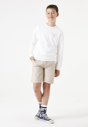 Sweatshirt blanc, shorts beiges et baskets montantes bleues associés à des chaussettes à rayures noires et blanches. Tenue décontractée avec des détails de design simples.