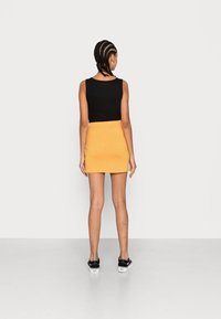 Una persona con el cabello trenzado lleva una camiseta sin mangas negra y una falda mini amarilla con un patrón, de pie de espaldas al espectador.