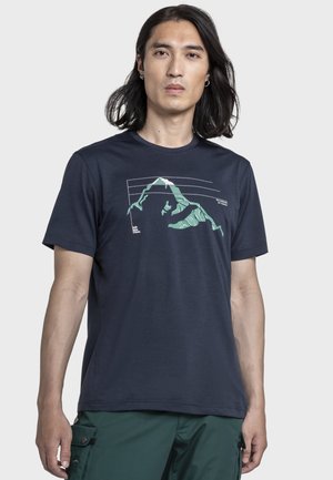 Mann mit schulterlangen dunklen Haaren, der ein marineblaues T-Shirt mit einer grünen Berggrafik und dunkelgrünen Cargohosen trägt.
