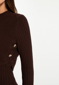 Maglione marrone a coste con collo alto, silhouette aderente e bottoni decorativi dorati sul lato. Tessuto morbido e strutturato.
