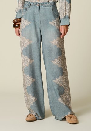 Hellblaue Jeans mit weitem Bein und beigen Spitzenmustern an den Seiten, getragen mit einer Spitzenbluse mit langen Ärmeln und hellbraunen Mokassins.