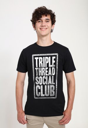Ung mand med krøllet hår, der smiler og har en sort t-shirt på med fed hvid tekst "TRIPLE THREAD SOCIAL CLUB" på forsiden.