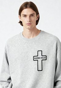 Jeune homme aux cheveux bruns raides portant un sweat-shirt gris clair avec une croix noire en contour et le texte « KARMA KILLS » sur la poitrine.