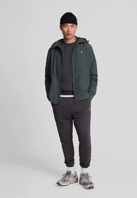 Tume roheline kapuutsiga jope halli dressipluusi kohal, sellega sobitatud tumedat halli spordipükstega. Omadused hõlmavad logodega aktsente, tekstuurset kangast ja tenniseid.