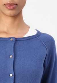 Μπλε πλεκτό πουλόβερ με στρογγυλή λαιμόκοψη, με κούμπωμα από κουμπιά και ριμπ άκρες. Μαλακή υφή με στενή εφαρμογή γύρω από τους ώμους.