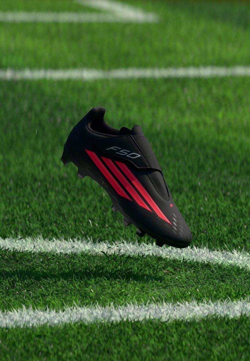Crampon de football Adidas F50 noir et rouge flottant au-dessus d’un terrain en herbe verte avec des lignes blanches de délimitation.