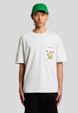 Ung mand i en hvid T-shirt med pink blomstret lomme-design og sorte bukser, iført en skriggrøn baseballkasket, der står mod en grå baggrund.