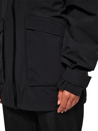 Rechte Hand einer Person, die eine schwarze Jacke mit großer Fronttasche und verstellbaren Manschettenriemen trägt.