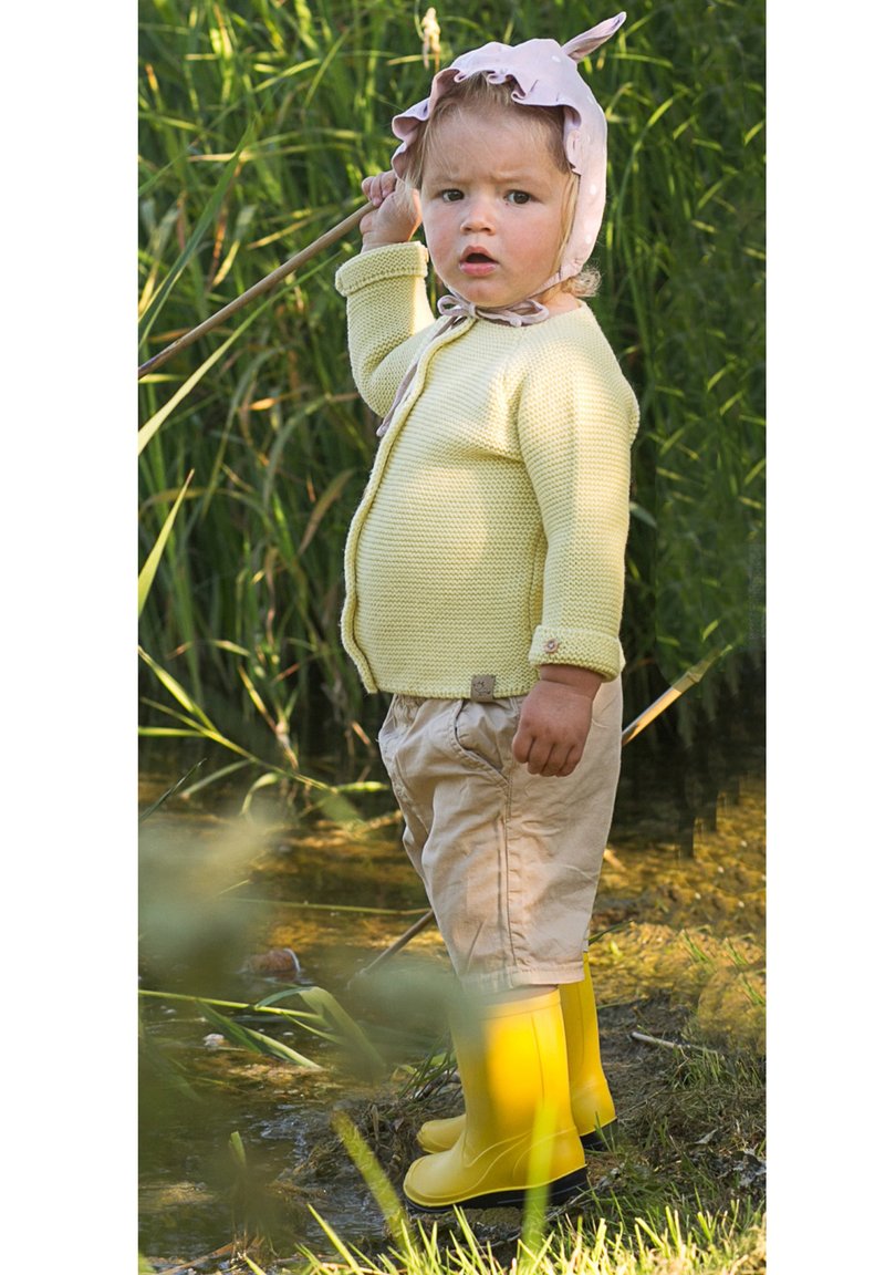 Kind in een lichtgele gebreide cardigan, beige shorts en felgele rubberen laarzen, staande in een grasrijk gebied naast water.