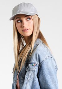 Sølvcap lavet af skinnende materiale, med en buet skygge og syede detaljer. Bæres sammen med en slidt denimjakke.