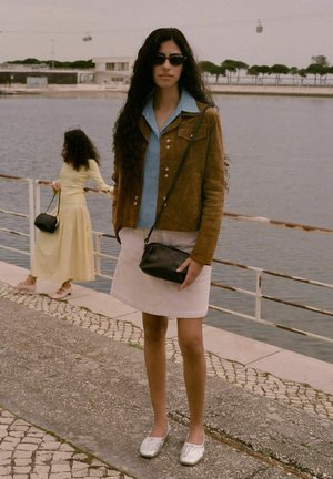 Giacca in suede marrone con camicia blu, gonna in corduroy bianca e ballerine argento. Borsa a tracolla nera e sfondo testurizzato con acqua.