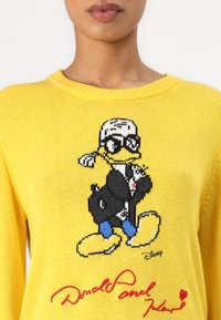 Pull en tricot jaune avec un design de canard en cartoon portant un manteau noir et des lunettes de soleil, agrémenté de texte rouge brodé en dessous.