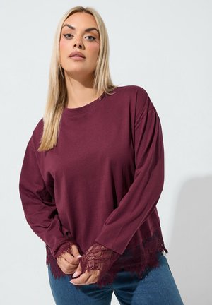 Jonge vrouw met lang blond haar die een bordeauxrode top met lange mouwen draagt, voorzien van kanten manchetten en zoom, gecombineerd met een blauwe spijkerbroek, poserend tegen een lichte achtergrond.