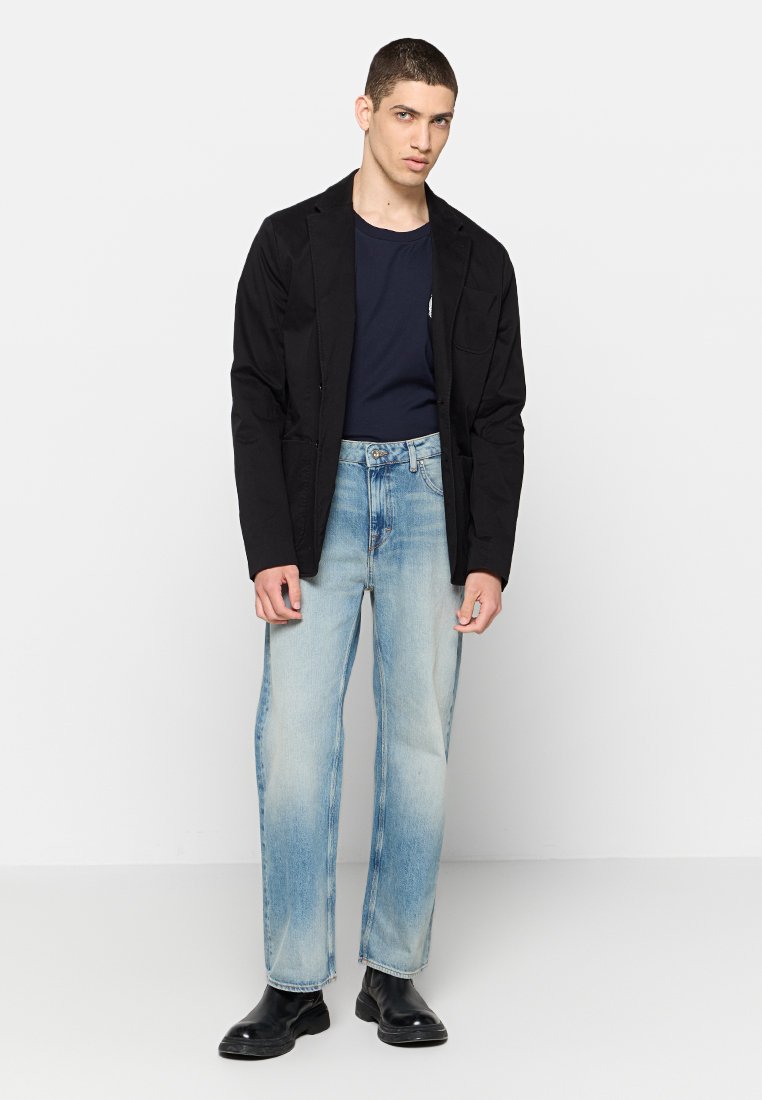 Schwarze Jacke mit strukturierter Oberfläche, marineblaues T-Shirt, hellblaue, stark ausgewaschene Jeans und schwarze Stiefeletten. Einfache, klare Linien, keine sichtbaren Muster.
