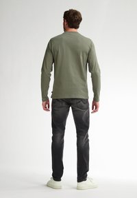 Cămașă verde măslin cu mâneci lungi și guler rotund, asortată cu blugi închisi și adidași albi, văzută din spate pe un fundal neutru.