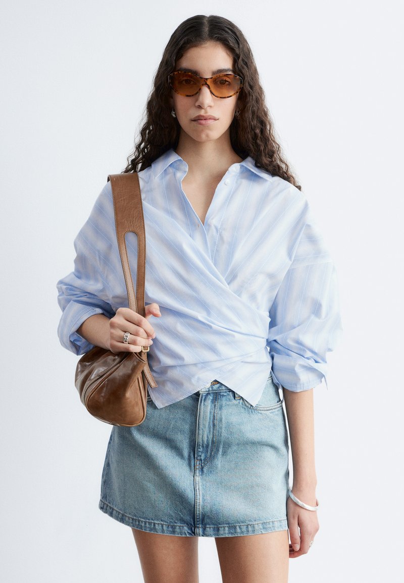 Femme aux longs cheveux bouclés portant des lunettes de soleil écaille, une chemise cache-cœur bleu clair, une jupe en jean, et un sac à bandoulière en cuir marron.